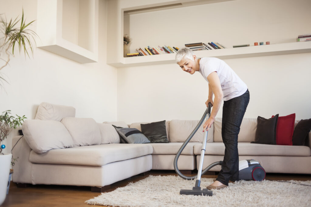 Woman vacuums a carpet in a living room with a large sectional sofa.