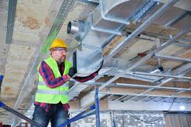 HVAC installer works on overhead metal ductwork in a construction area.